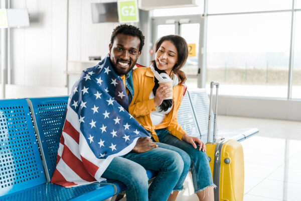happy african american couple sitting with american flag and baggage in departure lounge in airport